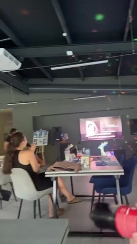 The video shows an indoor scene at what appears to be a cafe or restaurant with a dark ceiling and industrial-style exposed pipes. In the foreground, a young woman with long, dark, curly hair tied in a ponytail is seated at a table, wearing a black dress and holding a microphone. She is looking towards a large screen displaying text and images.

Another woman, wearing a black dress, is standing near the screen and gesturing. On the table in front of the seated woman are drinks and a bag. The screen displays text in Russian, including what appears to be a question or statement about love and beauty, and images of women. The setting is dimly lit, with colorful lights from a disco ball or similar fixture projected onto the ceiling, suggesting a lively or festive atmosphere. The overall mood seems to be engaged, with the seated woman actively participating in what looks like a presentation, karaoke, or a similar interactive event. The location context provided is Danilovka (Altai), Russia.
