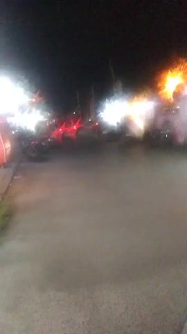 A blurry nighttime video shows a street scene with several vehicles, including motorcycles and possibly a truck. Bright lights from the vehicles and possibly storefronts illuminate the area. There are also some indistinct figures and objects in the background, suggesting a commercial or public space. The ground appears to be wet or reflective. The camera perspective is low, possibly from the viewpoint of someone on a motorcycle.