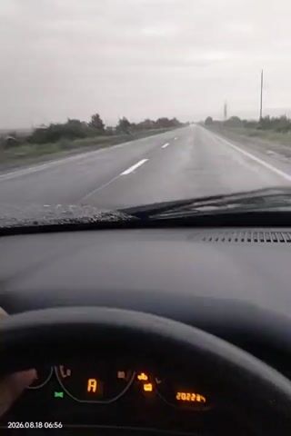 The video shows a first-person view from inside a car driving on a wet road under a cloudy sky. The windshield wipers are intermittently activated, clearing rain from the view. The dashboard displays a speedometer with the needle indicating a speed below 80, a gear indicator showing 'A' for automatic transmission, and an odometer reading of 202235. The date and time stamp in the corner reads 2026.08.18 06:56. The road is surrounded by vegetation on either side, and utility poles are visible in the distance. The overall atmosphere is overcast and appears to be during daylight hours.
