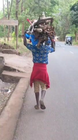 A person walks away from the camera, carrying a bundle of wood on their head. The person is wearing a blue patterned shirt, tan shorts, a red sweater tied around their waist, and sandals. They are walking on a paved road with a concrete sidewalk on the left. Trees are visible on both sides of the road. A three-wheeled vehicle is driving in the opposite direction on the right side of the road. A wooden sign is visible on the left. The person appears to be walking with purpose, their back straight, carrying the load with ease. The scene is typical of a rural or semi-rural environment.