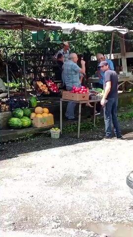 A roadside fruit stand is shown with several people present. On the left, there are watermelons and cantaloupes displayed in boxes. To the right, a table holds boxes of apples and what appear to be tomatoes. A man in a blue shirt is interacting with another man. In the background, a sign reads "ГОРЯЧАЯ КУКУРУЗА" (Hot Corn). A blue truck is partially visible in the foreground. The scene appears to be outdoors with trees and foliage in the background.
