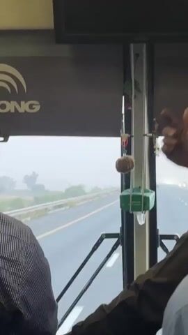 The video shows the interior of a bus looking out a window onto a highway. The bus is moving, and the road outside appears wet, suggesting rain or mist, making the distant landscape hazy. Trees are visible in the distance.

Inside the bus, a person wearing a striped shirt is seated with their back to the camera on the left. On the right, a person's arm and hand are visible, clad in a dark, long-sleeved garment. This person is gesturing with their index finger, pointing towards the window or the hanging decorations.

Various items are hanging from the bus window frame: a light brown, fuzzy, spherical ornament; a green rectangular object (possibly a small container or air freshener); and a set of keys. A black screen or monitor is mounted above the window. Part of a logo, "ONG", is visible on the bus interior panel on the top left, likely belonging to the bus manufacturer (e.g., YUTONG). The setting is a highway, potentially in Thatta Haji, Pakistan, given the provided context.