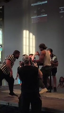 This is a video of an arm wrestling match. Two men are on either side of an arm wrestling table, gripping each other's hands. A referee in a striped shirt stands between them, observing the match. Another referee is to the right of the table, also watching. A person wearing a black shirt and a white baseball cap is filming the event from the front. A screen is projected on the wall behind the wrestlers, displaying information related to the match, including "TABLE 1" and "CURRENT RESULTS." The environment appears to be an indoor venue, possibly a hall or auditorium, with spectators visible in the background. The lighting suggests it is daytime, with sunlight casting shadows on the wall. The arm wrestlers are both focused on the match, with their bodies tensed. The referee on the left raises his arm at 00:04, indicating the end of the match.
