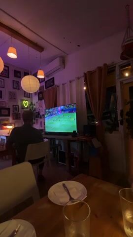 The video shows a dimly lit room with a television displaying a soccer game. A person is seated with their back to the camera, facing the television. The room appears to be a restaurant or cafe, with tables and chairs visible. There are several framed pictures on the wall, as well as decorative lighting. The overall atmosphere is relaxed and casual.