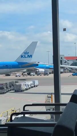 This is a short video of an airport scene viewed from inside. A large KLM airplane with its distinctive blue and white livery is parked on the tarmac. There are several other airplanes visible in the background, as well as airport vehicles and luggage carts. The sky is blue with some clouds. The video is shot from a seated position, and parts of seats and a window frame are visible in the foreground.