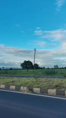 The video shows a view from a moving vehicle along a road. The road has a gray asphalt surface with a white and gray concrete barrier along the side. Beyond the barrier is a strip of green grass and vegetation. In the background, there are agricultural fields, trees, and utility poles with power lines stretching across the frame. The sky is blue with scattered white clouds. The overall scene depicts a rural or semi-rural landscape.