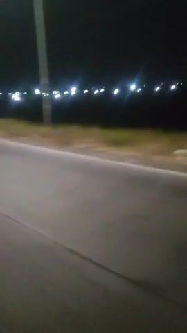 The video shows a nighttime scene from a vehicle moving along a road. The road surface is visible in the foreground, with a yellow-brownish embankment to the left and a line of bright white lights stretching across the horizon in the background. The lights appear to be streetlights or lights from a distant town, creating a blurred effect due to the motion of the vehicle and the darkness. The overall impression is of driving through a rural or suburban area at night.