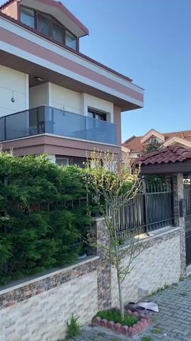 The camera pans from left to right, starting with a partial view of a two-story house with a light brown facade and a balcony. The house is surrounded by a low wall made of stone and a green hedge. Next to the wall is a small tree with sparse leaves. 

The pan continues to show more houses along the street, all with similar architectural styles and red tiled roofs. The street is paved with gray bricks and asphalt. 

In the distance, the sea is visible, stretching out to the horizon under a clear blue sky. Several cars are parked along the street, and a small group of people can be seen standing near the water.