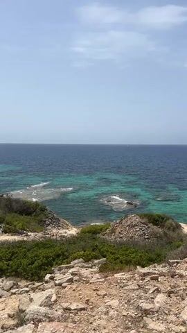 A vertical video shows a view of the sea from a rocky, vegetated cliff. The water is a deep blue in the distance, transitioning to a bright turquoise closer to shore, revealing submerged rocks and coral formations. The sky is mostly clear blue with scattered white clouds. The foreground consists of rough, light-colored rocks and patches of green shrubs. There are no people or visible man-made objects in the immediate scene, though a small white object, possibly a boat, can be seen on the horizon. The lighting suggests it is daytime.