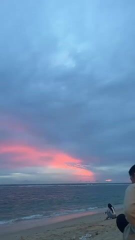 The video shows a beach scene with the ocean and a colorful sky. The sky is mostly gray with streaks of pink and orange clouds, suggesting either sunrise or sunset. The ocean is a deep blue-green with small waves rolling onto the sandy beach. In the foreground, on the right side of the frame, a person is partially visible, facing away from the camera and looking towards the ocean. On the beach, further to the left, a second person is sitting down, also looking towards the water. The beach appears to be composed of sand with some debris scattered on it. The overall atmosphere is calm and serene.
