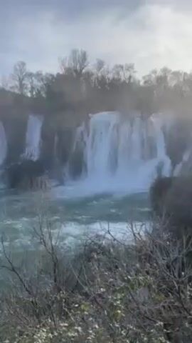 The video shows a waterfall cascading into a turquoise river. The water falls in multiple streams from different heights, creating a misty spray in the air. The surrounding landscape includes trees and shrubs, some of which are bare, suggesting it might be late fall or winter. The sky is bright, with some clouds visible. The camera slowly pans to the left, revealing more of the waterfall and the river it feeds into.