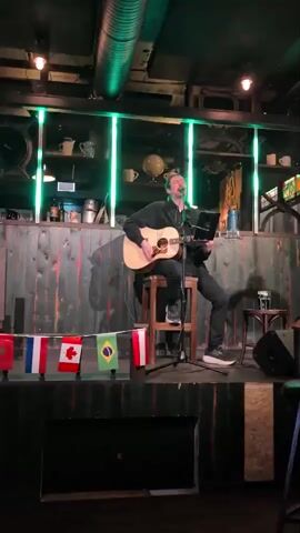 The video shows a man playing an acoustic guitar and singing on a small stage. He is sitting on a stool, with a microphone in front of him and a laptop on a stand to his right. The stage is decorated with a row of small national flags, including France, Belgium, Canada, and Brazil. Behind the stage, there is a bar area with shelves stocked with bottles and mugs. The lighting is dim, with green lights accentuating the bar area. The overall atmosphere appears to be that of a small music venue or bar.