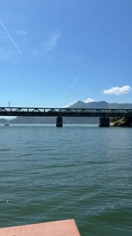 A static shot from a low vantage point on the water shows a wide body of water in the foreground and midground under a clear blue sky with a few contrails. A long, dark green metal truss bridge with multiple sections and concrete support piers spans the water in the midground, extending from left to right. Distant mountains with some hazy clouds are visible behind the bridge. A small boat is partially visible on the far left, near the horizon. A wooden edge, likely part of a boat or dock, is present in the bottom right corner of the frame.