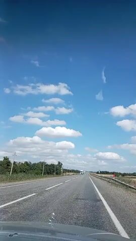 The video shows a first-person view of driving on a highway under a blue sky with scattered white clouds. The road is paved with asphalt and has white lane markings. On the left side of the road, there are trees and some utility poles. On the right side, there is a metal guardrail and more trees in the distance. Several vehicles are visible on the highway, including cars and trucks. The video pans slightly to the left and right, showing the passing scenery. The overall environment appears to be rural or semi-rural, with good visibility and clear weather.