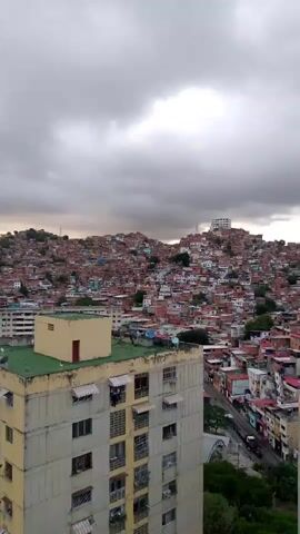 The video shows a panoramic view of a densely populated hillside with numerous buildings. In the foreground, there is a tall, light-colored apartment building with multiple windows and balconies. A lighter yellow structure with a green roof is also visible. The hillside is covered with a multitude of red-roofed houses, appearing to be a favela or shantytown. The sky is overcast with heavy, dark clouds. The overall scene depicts an urban environment characterized by both modern apartment buildings and informal settlements, under a cloudy sky. No people or specific actions are discernible in the video.