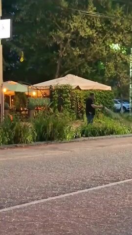 A man is watering plants in front of a restaurant with outdoor seating. A car drives by on the road in the foreground. The scene appears to be at dusk or in the evening, with lights visible from the restaurant.