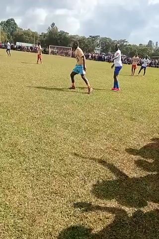 The video depicts a soccer game in progress on a grassy field. Players in blue and white uniforms are competing against players in gold and teal uniforms. A crowd of spectators lines the perimeter of the field. The camera follows the action of a player in a blue and white uniform with the number "9" on his back as he dribbles the ball. The sky is overcast.