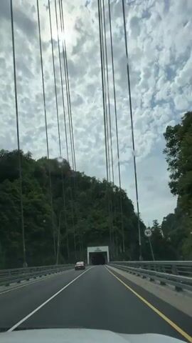 The video captures a point-of-view perspective from a vehicle traveling on a highway. The highway appears to be a bridge with tall cables descending from above, suggesting a suspension bridge. Dense green foliage covers the surrounding mountainside. Ahead, a large tunnel entrance is visible, framed by a white structure. A red car is seen on the highway to the left of the frame, and another red car is parked near the tunnel entrance on the right. The sky is filled with scattered white clouds, and the sun is partially visible, creating a bright spot. The road has clear lane markings.