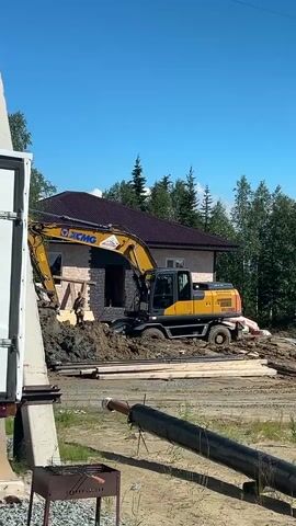 A yellow excavator is on a construction site. The excavator is positioned in front of a house that is under construction. There is a pile of dirt next to the excavator. A person wearing a dark shirt and light-colored pants is standing near the excavator. The excavator's arm is extended and appears to be digging into the dirt. There are wooden planks and pipes on the ground in front of the excavator. The sky is blue and clear. Trees are visible in the background. A portion of a white trailer or structure is on the left side of the frame. A charcoal grill is on the ground in the lower left corner.