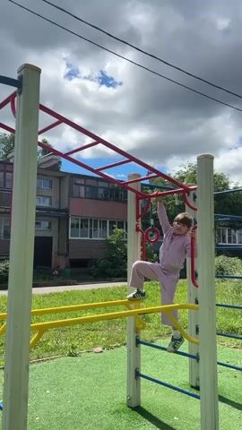 A young girl in a light purple tracksuit is climbing a monkey bar. She is on a playground with green turf flooring and yellow bars. Behind her is a residential building and a cloudy sky. She is actively moving along the monkey bar, demonstrating her physical coordination.