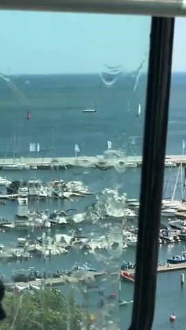The video shows a view from inside a vehicle, likely a tram or train, looking out a window. The window has water droplets and streaks on it, obscuring some of the view. The scene outside is a harbor filled with numerous boats of various sizes, including sailboats and motorboats, docked in the water. In the background, the calm sea stretches to the horizon under a clear, bright sky. A few small objects are visible on the water, possibly buoys or other vessels. The overall impression is a sunny day at a busy marina. The presence of boats and the sea suggests a coastal location. The style of the vehicles and the architecture visible in the distance (though not clearly detailed) are consistent with a European setting, and the mention of Gdynia, Poland, places the scene in a specific coastal city known for its maritime activities. The sunlight indicates it's daytime, likely late morning or afternoon. The water droplets on the window might indicate recent rain or condensation, but the clear sky suggests it's not currently raining. The mood is calm and observational, focusing on the tranquil scene of the harbor. There are no people clearly visible, but the presence of the boats implies human activity related to boating and maritime life. No text is clearly discernible on the window or in the distant view.