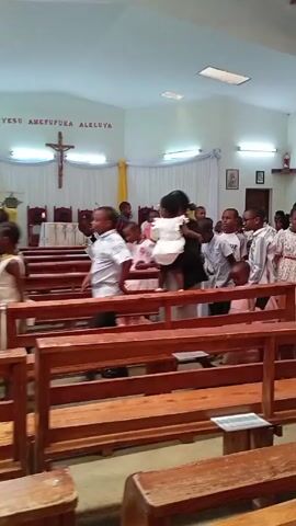 Here is a concise description of the video:

The video shows a group of children and some adults inside a church. The children are mostly wearing formal attire such as dresses and collared shirts, suggesting a special occasion. Some are standing near the wooden pews, while others walk around. Adults are interspersed among them, seemingly guiding or supervising the children.

The church interior features wooden pews, a cross hanging on the wall, and religious artwork or decorations. The space appears well-lit with natural light coming through windows. Overall, the scene seems to capture a moment of activity and movement within the church setting, possibly during or after a religious service.