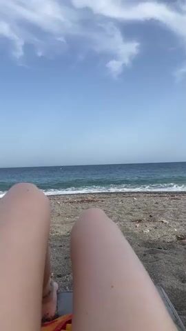 The video shows a person's legs stretched out on a beach chair, facing the ocean. The sun is shining and the sky is blue with some white clouds. The ocean waves are gently breaking on the shore. There are some small pieces of seaweed or debris on the sand near the water. The person's feet are partially visible at the bottom left of the frame. The overall impression is one of relaxation and enjoyment of a beach day.