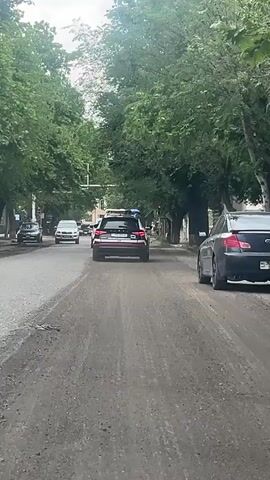 The video captures a daytime street scene in Chiva, Armenia. A black police SUV with active red and blue flashing emergency lights is driving away from the camera. Its license plate is partially visible, with "707" discernible.

Other vehicles are present on the road: a silver SUV is approaching in the opposite lane, a black sedan is parked on the right side of the frame, and several other cars are visible further down the street. The road surface is worn asphalt, with a lighter, possibly dusty or gravelly median strip.

Both sides of the street are lined with mature trees featuring dense green foliage, with glimpses of buildings visible behind them. A red traffic light is visible in the distance. The scene depicts an urban or suburban street with traffic, focused on the moving police vehicle.