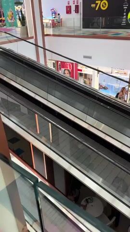 This media file captures a view of a multi-level shopping mall, likely in Adeje, Spain. The scene is brightly lit, suggesting it's daytime. The primary focus is on the escalators, which are moving. Several people are visible on different levels of the mall, some walking, and others appearing to be seated at small tables, possibly in a food court area.

Decorative elements in the form of large, inflated soccer balls are suspended from the ceiling at various points in the mall. These soccer balls are bright yellow and blue, and some are also colored red and blue. They add a playful and possibly festive atmosphere to the environment.

On the upper levels, storefronts with various merchandise are visible. One sign displays "70," and another large banner reads "SURE AL MUNDO DE LOS JUGUETES" (Welcome to the World of Toys). Other smaller signs with text are also present but are not easily decipherable. The architecture of the mall features modern design elements with metallic finishes and warm lighting.

The overall impression is of a typical shopping mall environment, bustling with activity and decorated for a special event or promotion. The presence of people and the functioning escalators indicate normal operations. The soccer ball decorations suggest a theme, perhaps related to a sporting event or a children's activity.