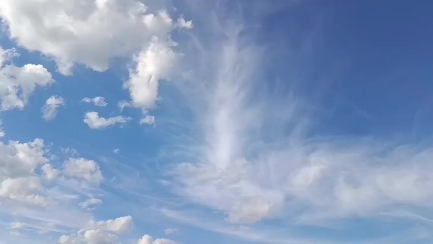 This is a video of a bright blue sky with scattered clouds. The clouds are mostly white and fluffy, with some wispy cirrus clouds also visible. The sun is shining brightly, suggesting it is daytime. There are no people or specific objects clearly visible in the frame, but the clouds and sky create a serene and expansive natural scene. The video appears to be a static shot of the sky, with no discernible movement other than the subtle shifts in the clouds. Given the location context of Lahojsk, Belarus, it depicts a clear and pleasant day in a rural or natural setting. The overall mood is peaceful and uplifting. There is no visible text in the media file.