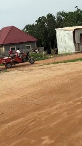 The video begins by showing a dirt road with a red motorized tricycle with a trailer parked on the left side. Two men are near the tricycle, one appearing to be getting into the trailer. Behind the tricycle, there is a small, corrugated metal structure resembling a shop, with a wooden counter in front of it. A second, similar corrugated metal structure is situated to the right of the first one. Green trees and foliage are visible in the background. In the following frames, the tricycle begins to move forward and slightly to the right. One of the men is now in the trailer, and another is in the driver's seat. The tricycle continues to move, and the scene shifts slightly, revealing more of the surrounding environment, including another building with a red roof to the left of the shops. The video ends with the tricycle continuing to move forward.