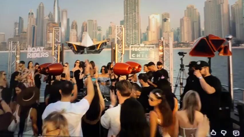 The video shows a large group of people on a boat with a city skyline in the background, likely Dubai. The boat appears to be hosting a party or event, with a DJ setup and speakers visible. Many people are standing and socializing, some are holding up phones, and one person is wearing a straw hat. The overall atmosphere seems lively and festive.