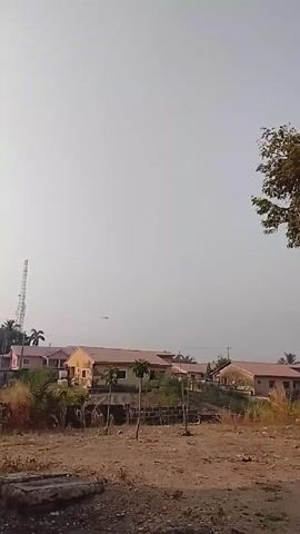 The video shows a residential area with single-story houses. The houses are painted in light colors with red tile roofs. A cell tower is visible in the distance to the left. In the foreground is a dirt area with some sparse vegetation. Above the houses, a helicopter flies across the sky. The sky is overcast and pale. A large tree is visible on the right side of the frame.