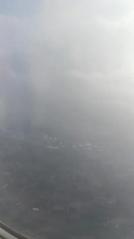 This video shows an aerial view from an airplane window, looking down at a landscape partially obscured by clouds. The ground below appears to be a mix of agricultural fields and some buildings, possibly a small town or rural area. In the distance, there is a large, complex structure that resembles a theme park or a large modern building with a distinctive architectural design, perhaps with towers or arches. The sky is partly cloudy, with sunlight filtering through, creating a hazy effect. The perspective suggests the airplane is either ascending or descending, as the view is angled downwards towards the earth. The image quality is somewhat blurry, likely due to the airplane window and motion.