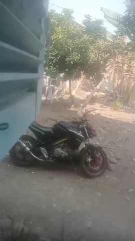 A black motorcycle with a red rim is parked on a dirt path next to a building with blue siding. Trees and vegetation are visible in the background. The scene is illuminated by daylight, suggesting it is outdoors.