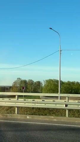 The video shows a view from a vehicle moving along a road or highway. The sky is blue with some faint clouds. A metal guardrail separates the road from the adjacent landscape, which consists of green fields, trees, and some distant buildings. There are tall, white lampposts and utility lines. A road sign with arrows indicating direction is visible at the beginning of the video. The video moves steadily forward.