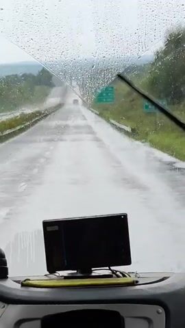 A forward-facing view from inside a vehicle on a wet, overcast day. Raindrops are visible on the windshield, and the road ahead is slick and appears to be partially flooded. Green signs with text are visible on the right side of the road. A black electronic device is mounted on the dashboard in the foreground, with a black hemisphere object to its left. The road extends into the distance, with a few vehicles visible further ahead. Trees and hills line the sides of the road.