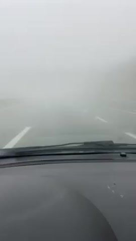 The video shows the view from inside a car, looking out through the windshield. The car is driving on a road with white lane markings. The weather is very foggy, severely limiting visibility. Only a short distance of the road ahead is visible through the fog. The dashboard of the car is visible at the bottom of the frame. A bridge or overpass briefly appears and disappears in the distance due to the fog.