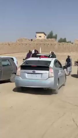 The video shows a dusty outdoor scene.  A light blue Prius is parked on dirt, with several men standing on and around it.  Some of the men are wearing turbans. A white flag is visible on the car. A brick wall and a small building are visible in the background. There are two other cars parked nearby, one gray and one white.  The sky is clear and blue.