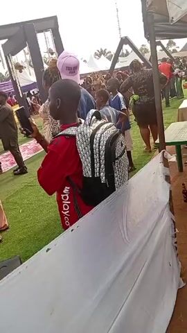 The video shows a crowd of people gathered at an outdoor event, likely a party or celebration, on what appears to be artificial turf.  In the foreground, a young person wearing a red shirt and a Dior backpack is taking photos with a phone.  Scattered around the base of a large black speaker are numerous paper bills.  People are interacting with the money, picking it up and distributing it.  There is a tent-like structure nearby, along with tables and other event decorations.  The overall atmosphere suggests a lively, festive occasion.