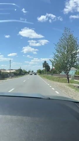 The video shows a view from inside a vehicle looking out at a rural road on a sunny day. The sky is blue with scattered white clouds. There is a paved road with a white line marking the lane. On the left side of the road, there are houses, trees, and a utility pole. On the right side of the road, there is a grassy area with a fence, trees, and utility poles. Several vehicles are visible on the road, including a white van, a truck, and a few cars. The truck is white and has a trailer. The van is also white. The vehicles are moving away from the camera. The overall impression is a typical scene of driving through a rural area.