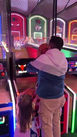 This vertical shot captures an active scene inside an arcade in Columbus, United States of America, featuring multiple basketball shooting games. In the center, an adult male, wearing glasses, a two-tone hoodie (blue, white, and grey), and tan cargo pants, is playing an arcade basketball game. He is shown mid-action, with a basketball in the air, and then turns to look at the game screen, which displays "GAME OVER," a score of "23," and other text like "SCORE," "BONUS," and "PRESS COMP."

To his left, partially visible in the foreground, a small child with long dark hair, wearing a light-colored top, holds a basketball, appearing to be either participating or observing the game. The arcade is brightly illuminated by vibrant neon lights, with blue lights on the left game unit, green on the right, and red accents on the central machines. The games feature "NBA Hoops" branding and prominent "CHICAGO BULLS" logos on the backboards. The overall setting is lively and recreational.