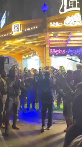 The video shows a group of men dancing and celebrating outdoors in front of a shop with Arabic signage. The shop has a wooden facade adorned with lights. One man, dressed in a unique patterned jacket, sings into a microphone while others clap and cheer. A person in the crowd records the performance on their smartphone. A black cloth is waved in the air. The atmosphere appears festive and lively.