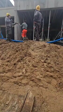 Three construction workers are visible. The worker on the left is wearing a yellow hard hat and is holding a rope. The worker in the center is wearing an orange pair of pants and is pulling on the same rope. The worker on the right is wearing a yellow hard hat and is standing and watching. The workers are on a dirt mound in front of a concrete structure. There are scaffolding and blue hoses visible in the background. The weather appears to be overcast.