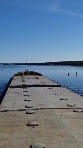 The image presents an elevated view looking forward over the deck of a large vessel, likely a barge or cargo ship, as it navigates a wide, calm body of water. The expansive, flat, gray deck dominates the foreground, featuring multiple rows of circular hatch covers. A low railing runs along the sides of the deck. Towards the far end of the vessel, a small superstructure or wheelhouse with antennae is visible.

The water ahead is a tranquil, deep blue, perfectly reflecting the clear, cloudless sky above. Slight ripples on the water's surface to the left suggest the vessel is in motion. The distant banks of the waterway are covered in dense green trees, indicating a natural or forested landscape. On the right side of the water, a prominent red navigational buoy stands vertically, casting a clear reflection on the still surface. Faint vertical structures are barely visible on the distant horizon against the bright sky.

The scene is illuminated by bright daylight, suggesting either morning or early afternoon. The weather is clear and sunny, contributing to the serene and open atmosphere of the image. The setting appears to be a wide river or a large lake. There is no visible text in the image. Given the provided context, the location is Kosino, Russia.