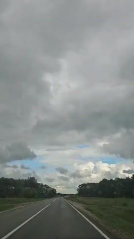 This is a first-person perspective video of driving on a paved road surrounded by trees under a cloudy sky. The road is gray with white lane markings. The sky is filled with heavy gray clouds with some patches of blue sky visible. The trees on either side of the road are lush and green. The video captures the feeling of a journey on a slightly overcast day.