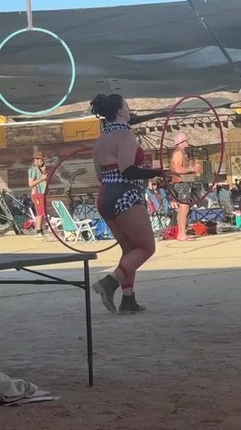 The video shows a woman in a distinctive harlequin outfit, consisting of a black and white checkered top and matching shorts with red accents, along with red fishnet stockings and black boots. She is holding two red hula hoops in her hands and appears to be preparing to use them. The setting seems to be an outdoor event or gathering, with a dusty ground and various makeshift structures and canopies visible in the background. People are milling about, some sitting in chairs. One man is wearing a hat and patterned shirt, while another walks past in a white shirt. A table is partially visible in the foreground. A cyan hoop is suspended from the canopy structure above. The scene has a somewhat festive or unconventional atmosphere.