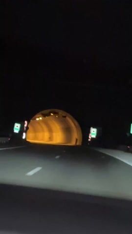 The video shows a blurry, low-angle view of a car approaching and entering a tunnel at night. The tunnel is illuminated with yellow lights, revealing its rounded shape and concrete structure. Road signs are visible on the sides of the road leading to the tunnel entrance. The camera appears to be mounted inside the car, capturing the perspective of the driver. The music playing in the background is audible.