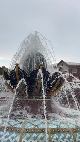 A fountain sprays water into the air. The fountain is made of stone and is decorated with mosaics. The water sprays upwards and then falls back into the fountain's basin. The fountain is located outdoors, with a building in the background. The sky is overcast. The video is shot from a low angle, looking up at the fountain.