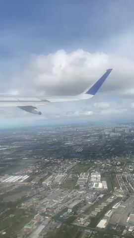 The video shows an aerial view from an airplane window. The airplane wing, painted white with a blue tip, is visible in the foreground. Below, the landscape consists of a sprawling urban area interspersed with green spaces, likely vegetation or farmland. The sky is partly cloudy, with patches of blue visible between the clouds. The overall lighting suggests a bright, sunny day. The plane appears to be in flight, descending or ascending over a city.